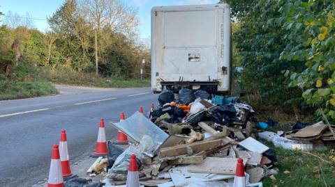 The front view of a white lorry trailer. In front of it are black plastic bags full of waste, wood offcuts and other waste which have been coned of with orange and white cones. They are on a lay-by beside a road