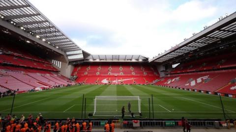 A general view of Anfield Stadium in Liverpool. The red seats are empty and a group of people in orange vests are walking in front of the pitch. 