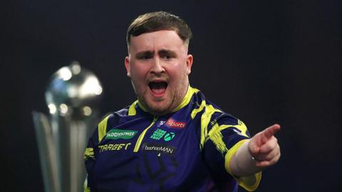 Luke Littler points with his outstretched left hand during the World Championship final. He has dark hair brushed to his right and is wearing a purple shirt with yellow trim. It has several sponsor logos including Sky Sports and Paddy Power. The world championship trophy can be seen over Littler's shoulder.