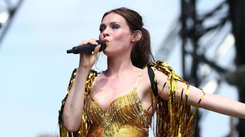 Sophie Ellis-Bextor wearing a gold outfit on stage, covered in gold streamers. She is singing into a microphone and has an outstretched arm.