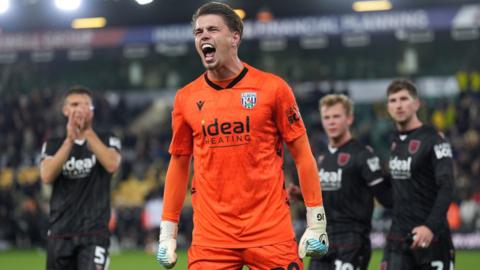 goalkeeper josh griffiths balls his gloved hands into fists and opens his mouth wide in a shout, his neck straining. he wears a bright orange shirt and has three teammates behind him.