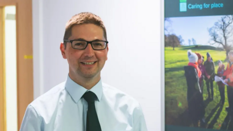 Head and shoulders shot of Richard Stanley, wearing glasses, a white shirt and black tie looking at the camera and smiling. The image is taken in an office corridor with a banner about caring visible in the background.