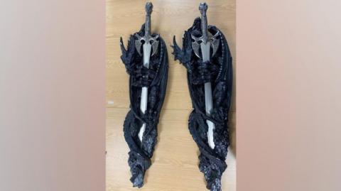 Two knives with elaborate handles stored in a dark decorative frame, held in place by a dragon-like carving.