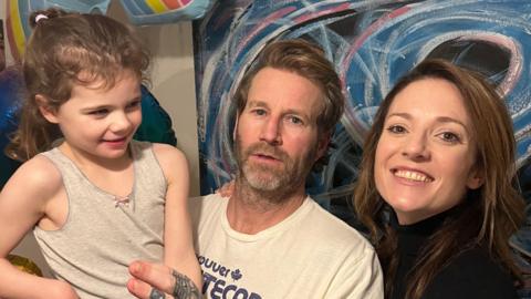 A man and a woman stand together while the man holds their young daughter. The man is wearing a white T-shirt and has a dark beard. The woman is smiling. She has long dark hair and is wearing a black top. The girl is looking at the woman. She is wearing a light grey vest top and has curly light brown hair.