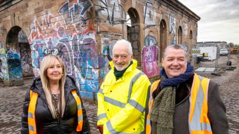 Deirdre Gaughan,  Andy Milne and Councillor Ricky Bell stand in front of the pump house. The building is covered in graffiti. 