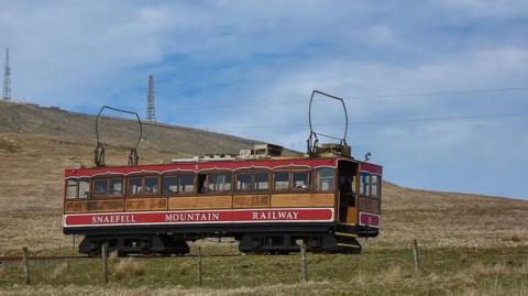 A a red and brown tram with the words Snaefell Mountain Railway in white travelling on tracks on a  green hillside. There is a blue sky above and two aerial masts can be seen on Snaefell in the background.