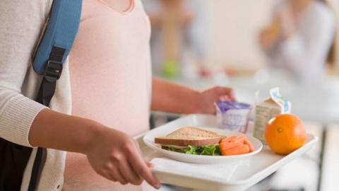 Child carrying tray of school canteen food including sandwich and orange