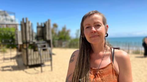 Kat stands in the play area. You can see play equipment behind her, and sand on the ground. Kat is wearing an orange dress and has long brown dreadlock hair. 