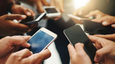 Six smartphones are held by people facing in to a circle, we can only see their hands and those at the back of the shot are slightly out of focus.