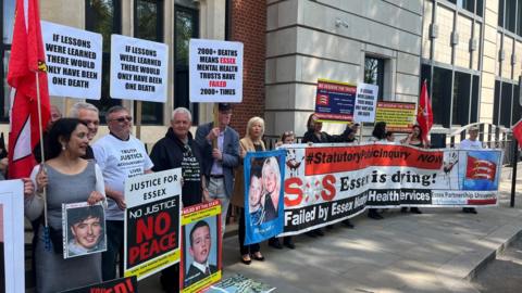 Protesters stand outside Arundel House in Central London. They are holding a big horizontal banner that says "SOS Essex is dying!" Some are also holding posters of their relatives that have died. They are standing outside the front of the mock-Tudor building. There is a row of about 30 people.