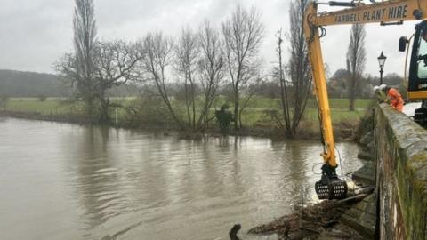 Workers looking over stone bridge with yellow arm of machinery above tree and debris under bridge in river