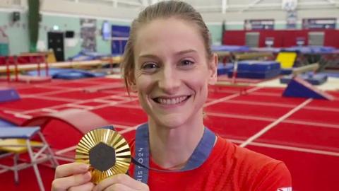 A smiling Bryony Page posing with her Paris 2024 Olympic gold medal. She has fair hair which is tied back. She is wearing a red Team GB top and is standing in a large sports hall with a red floor. Behind her are a number of blue trampolines and ramps.