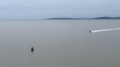 People stand huddled together, knee deep in water, as a speedboat races towards them.