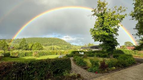 A rainbow arcs from left to right with a hedge and tree in the foreground and fields and mountains behind