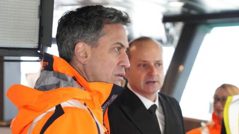  Ed Miliband is on board a ship wearing an orange jacket. Next to him is a man wearing a black jacket and tie, and a white shirt. A woman's face can be seen, blurred, to the right, in the background