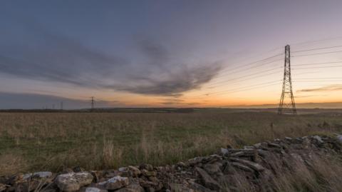 A photo showing a field with a number of pylons stretching across the landscape. There is a wall at the front and the skies are partly grey with sunshine in the distance