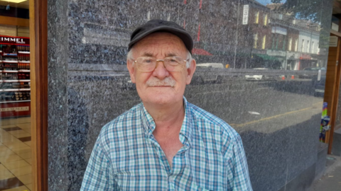 A man with white hair and a mustache and clear glasses looks into the camera. He is wearing a dark coloured hat and and open-neck blue check shirt.