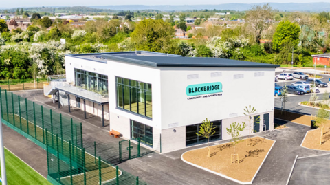 Blackbridge Community Sports Hub in Gloucester. It is a new white building with large windows and a balcony on the side, overlooking a pitch. There is fresh landscaping surrounding the building and trees and homes in the distance. 