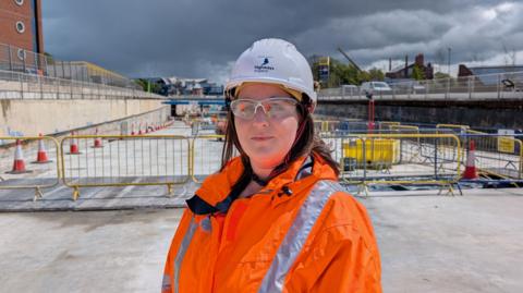 A woman wearing a bright orange hi-vis jacket and white safety helmet, She is standing in an area that is under the level of a main road with cones and barriers in the background.