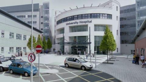 An exterior shot of University Hospital Coventry. Above the white round entrance building a sign reads "University Hospital". Cars are driving along a road in front of the building and people are walking on a pedestrianised area.