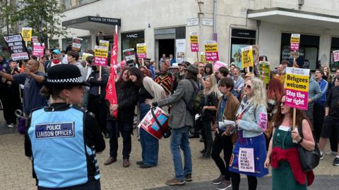 A crowd of people holding signs and looking to the left towards the counter protesters. There is a police liaison officer stood in front of the crowd wearing a blue jacket and a hat.