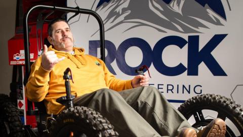 A man in a yellow hoodie sits in a large mobility chair with his hand up next to a white "Rock Engineering" sign.