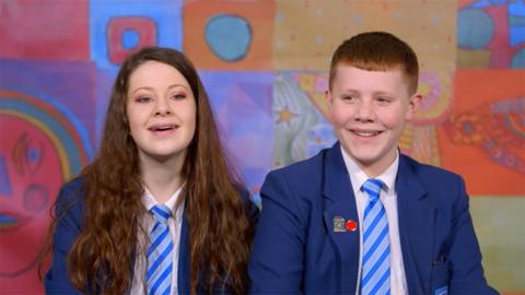 Photograph of schoolgirl and schoolboy smiling in front of a colourful classroom backdrop.