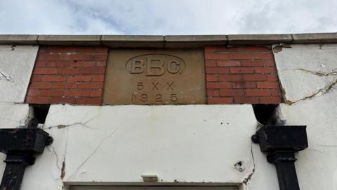 Detail from a brick building, showing the area above the door. There is exposed brick either side of an engraved stone which reads "BBC 5XX 1922". There is white plaster below and to the sides of the brick, alongside two black drainage pipes.