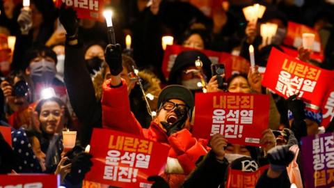 A crowd of protesters fills the frame, holding electric candles and red placards. One man in focus in the centre holds a gloved hand to his mouth as he yells