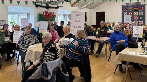 People sat around tables in a wooden-floored hall, wearing coats and holding white placards with slogans including Save our Service and We live in Hope