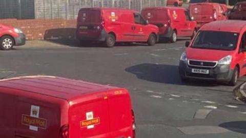 Eight red Royal Mail Vans are parked on roads either side of a T junction. There's a short brick wall with wooden fence above it in the background with a pavement in front of it.
