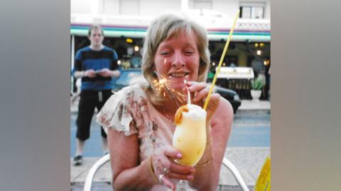 Annette Smith sitting at a cafe outdoors, drinking a yellow cocktail through a straw - it has a firework in one side. She appears younger, maybe in her 50s or 60s, and is wearing a cream sleeveless top with leaf patterns on. A man is standing in the background