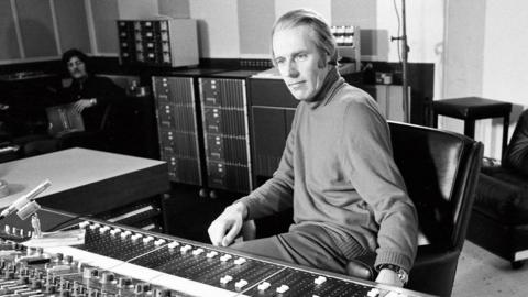 A black and white photograph of George Martin sitting at a mixing desk in a recording studio in 1971. 
