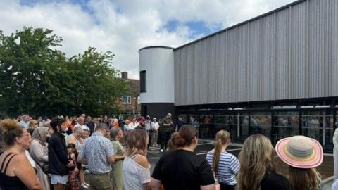 The building is made of glass panels on the ground floor, and is cladded in grey above. The picture is taken outside, where members of the public have gathered for the opening.