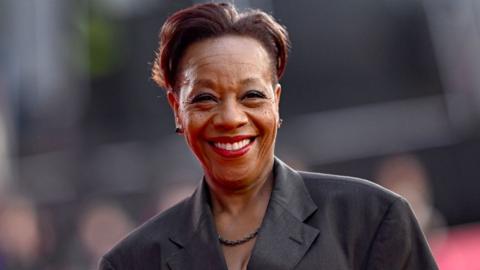 A grinning Marianne Jean-Baptiste looks at the camera as she wears a black suit jacket, black necklace and earrings