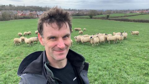 A farmer is standing in a green field with his flock of sheep behind him. Beyond the field, you can see houses. He is wearing a coat and black t-shirt.