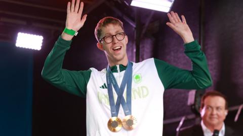 Daniel Wiffen on stage in Dublin at Team Ireland's homecoming ceremony from the Olympic Games. He is wearing his Team Ireland top and has his gold and bronze medals around his neck.