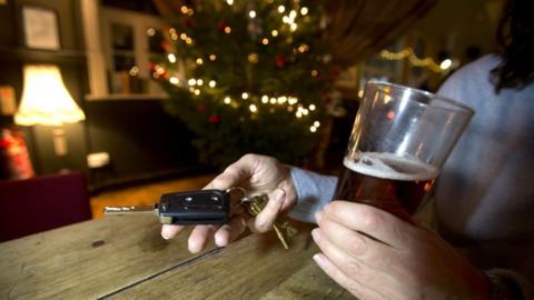 A pair of woman's hands holding an alcoholic drink in one, and car keys in the other with a lit up Christmas tree in the background.