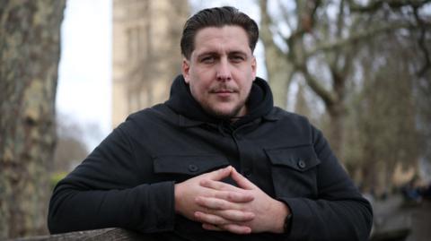 A man in a black jacket is looking at the camera. He has brown hair and has his hands clasped in front of him. Behind him are trees