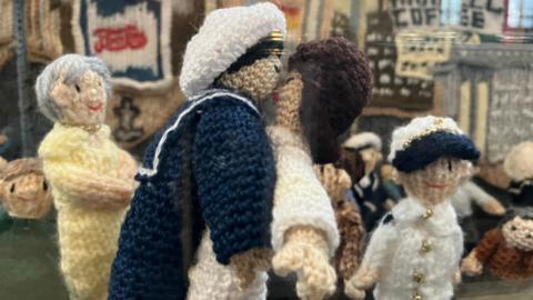 Knitted figures replicating a scene from World War Two. It shows a sailor in navy uniform dancing with a woman in a white dress. Behind it is a grey-haired woman in a yellow dress who is smiling, and a child in white and blue who is also smiling.