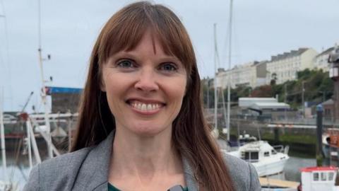 Joney Faragher, smiling: she has long straight brown hair with a fringe. She wears a green top under a grey jacket and stands in front of the quay in Douglas, with boats pictured behind her.
