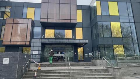 The front entrance of County Hall in Trowbridge with a person wearing a white hat and green trousers walking towards the entrance.