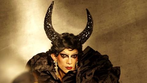 Zeeshan Ali in drag, wearing black horns and a short black wig