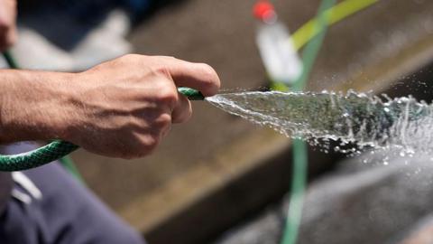 A close-up picture of a hand using a green hosepipe. Water is spraying out of the pipe.