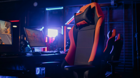 A large cushioned gaming chair, that is black and orange. There are two chairs behind in the background in a row, with a row of computer monitors in front. It is a dimly lit room. 