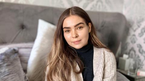 A young white woman with brown eyes and long brown hair sits on a grey bed with cushions around her. She's wearing a beige cardigan and smiling.