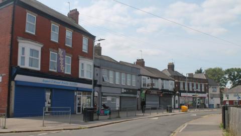 A road in Hull showing a row of high street shops with shutters closed. There is a path and a road in front of the shops. The street is empty and the sky is cloudy.