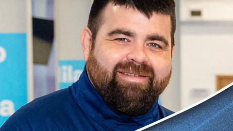 A head-and-shoulders image of a bearded man smiling at the camera. He is wearing a blue high-neck jumper.