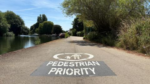 A tow path with a large white 'PEDESTRIAN PRIORITY' sign painted in large white letters on the path. The sign includes a circle with a parent and child painted in the centre. In the background, you can see boats moored.