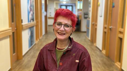 Fiona Carey pictured in a hospital ward's corridor. She is wearing a burgundy jacket, a close colour match to her short red hair and lipstick. She also wears a khaki green top, gold necklace and oval framed glasses. She is smiling at the camera. The corridor has wooden doors, white walls and brown laminated flooring.